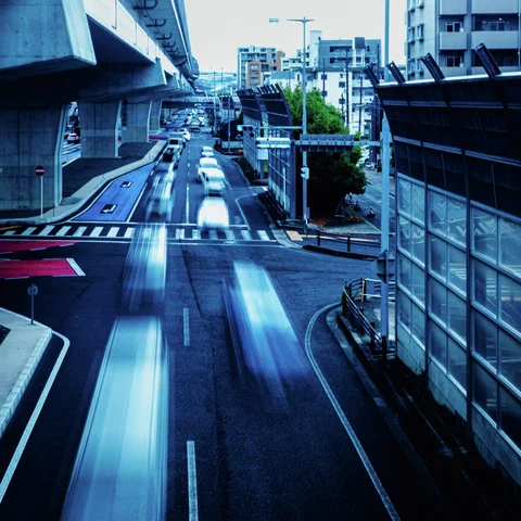 Long blue-white vehicle trails stretch along a city road beneath an elevated structure and tall barriers.