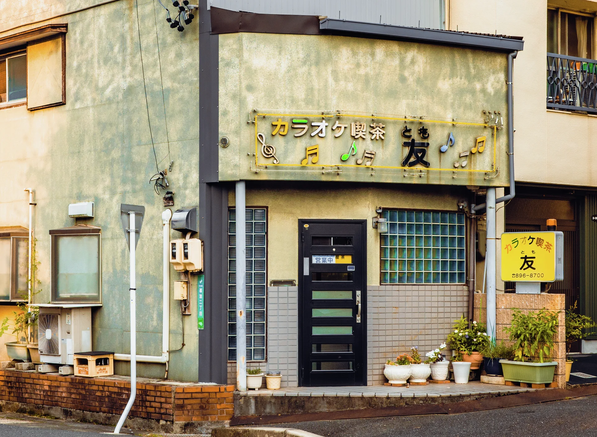 A weathered corner storefront has a black door, barred window, musical sign, and potted plants.