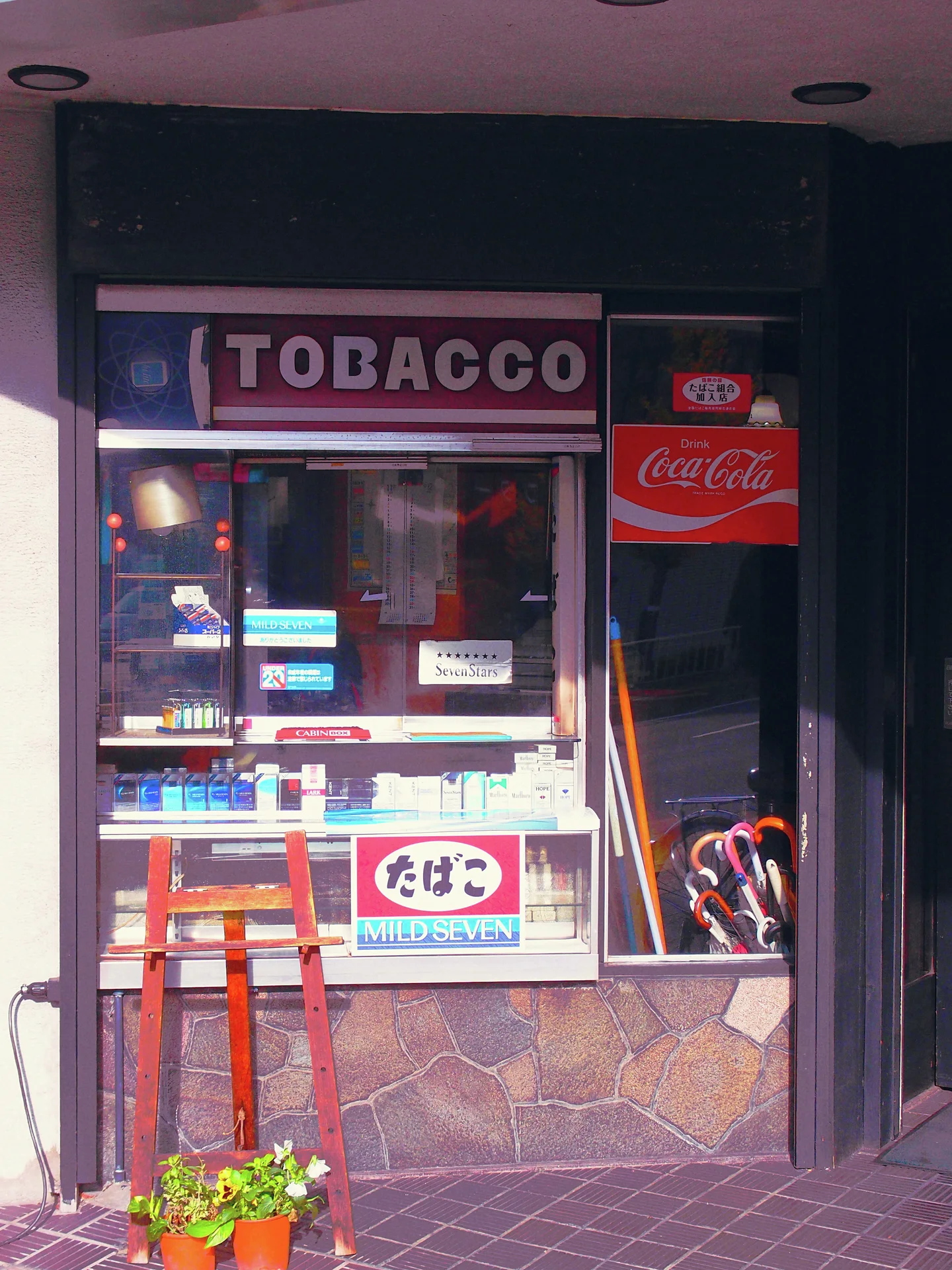 A small tobacco storefront has a glass display, red stool, and umbrellas beside its narrow entrance.