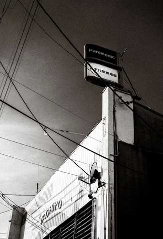 Utility wires crisscross above a narrow building topped by a projecting Panasonic sign in black and white.