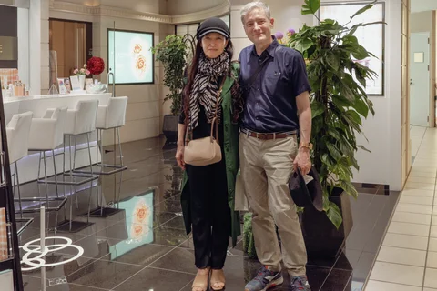 Two adults stand together beside a large leafy plant in a bright interior with white counter stools.