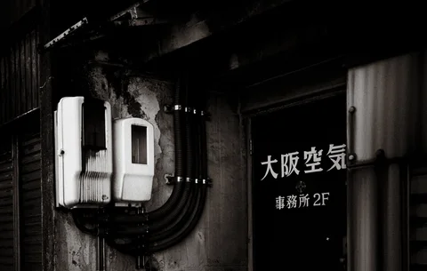 Two pale electrical boxes and curved black conduits sit beside a dark lettered door.