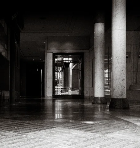 Tall columns border a dim polished hall leading to a distant doorway in a black and white view.