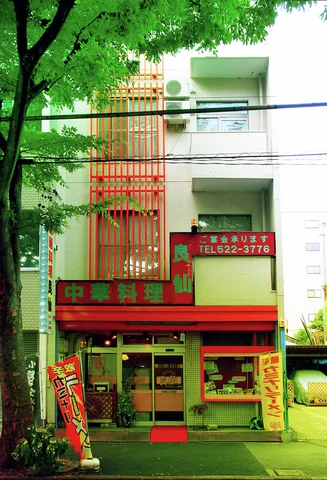 Red vertical slats and bright shop signage mark a narrow pale building beneath a leafy tree.