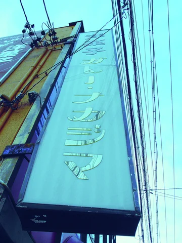 A tall blue-toned sign projects beside a building under dense overhead wires.