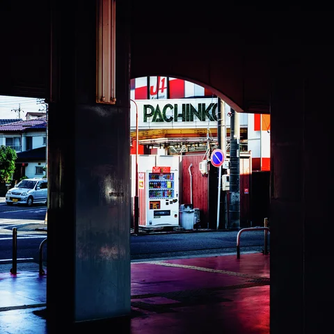 A shadowed concrete passage frames a bright red-and-white storefront and vending machine across the street.