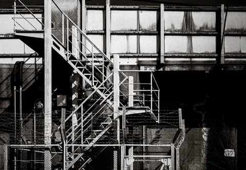 An exterior metal staircase zigzags across a weathered industrial wall behind fencing in monochrome.