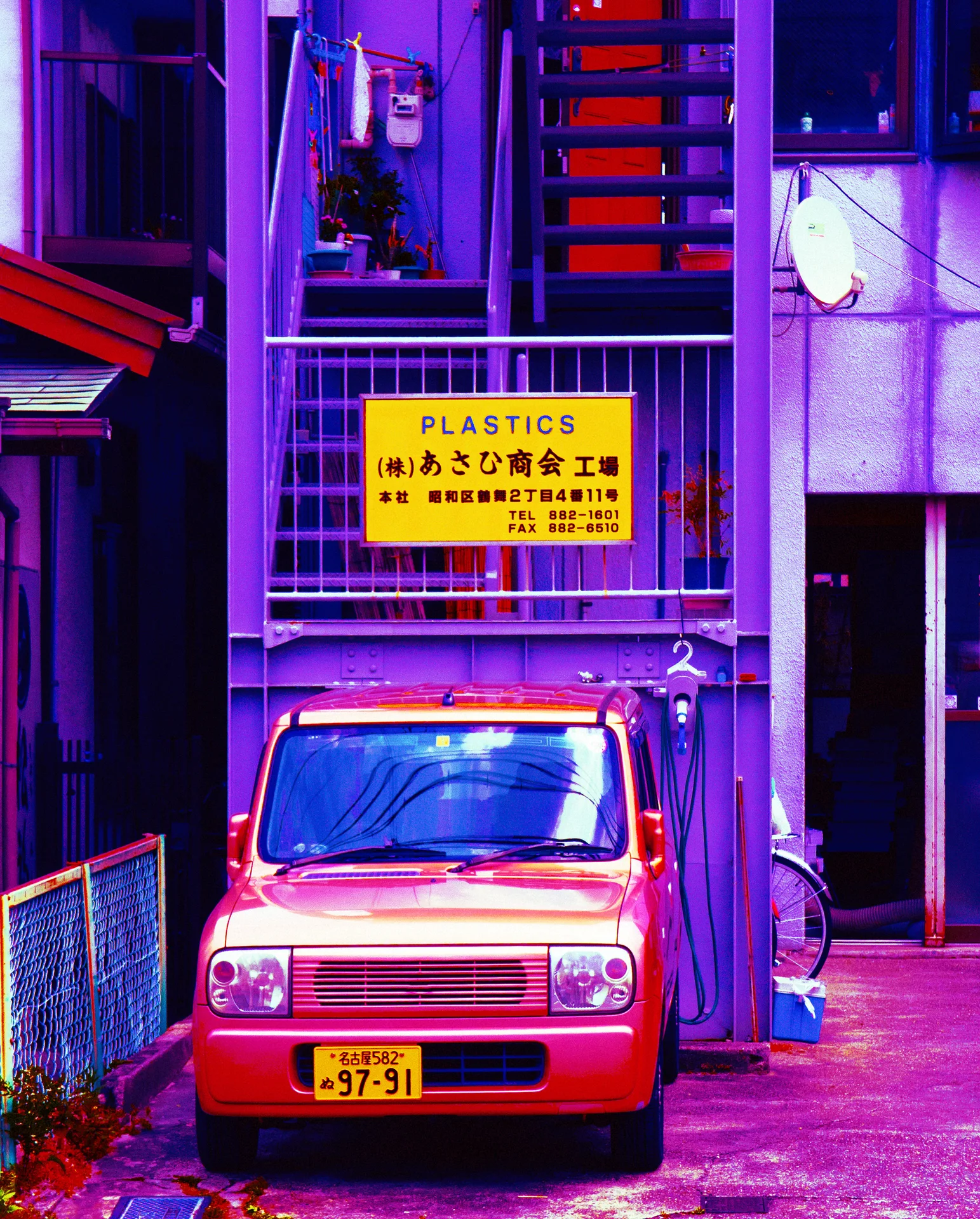 A small red vehicle sits beneath an exterior staircase and a yellow PLASTICS sign.