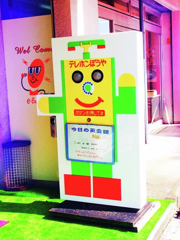 A colorful robot-shaped sign stands outside a glass-fronted shop on green matting.