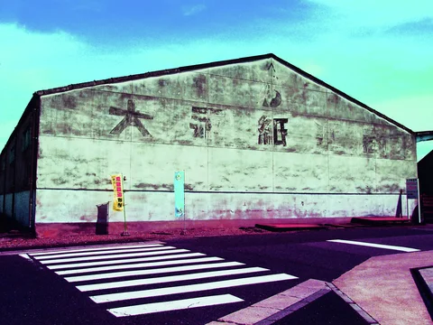 A broad weathered warehouse wall faces a white-striped pedestrian crossing beneath turquoise sky.