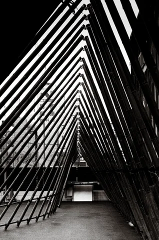Repeated diagonal metal braces form a long triangular corridor beside a building in black and white.