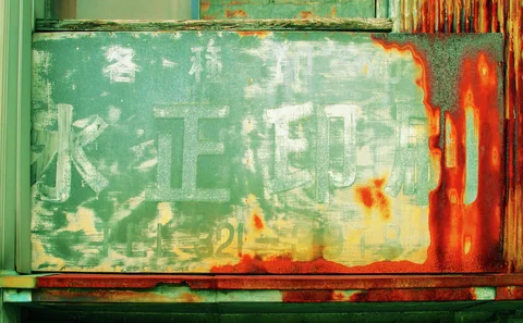 Faded Japanese lettering covers a green metal sign streaked with bright orange rust beside corrugated siding.
