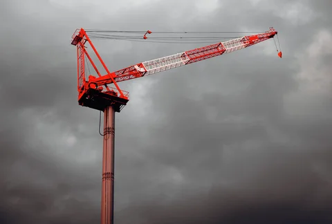A red and white tower crane stretches across a dark cloudy sky.