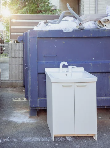 A white sink cabinet stands in front of a purple dumpster piled with discarded materials.