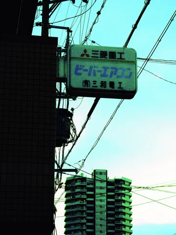 A small pale blue projecting sign hangs above a shadowed wall beside utility wires.