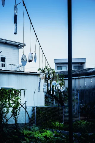 Small hanging containers dangle from overhead lines between low buildings above garden greenery.