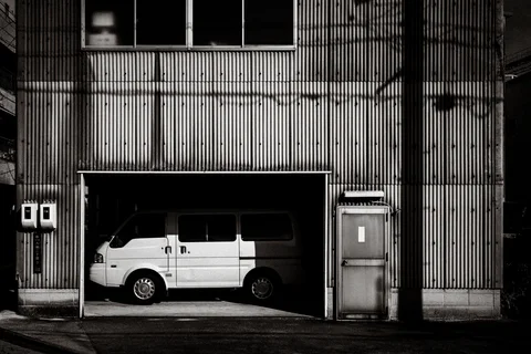 A white van sits inside an open garage bay in a corrugated building in black and white.