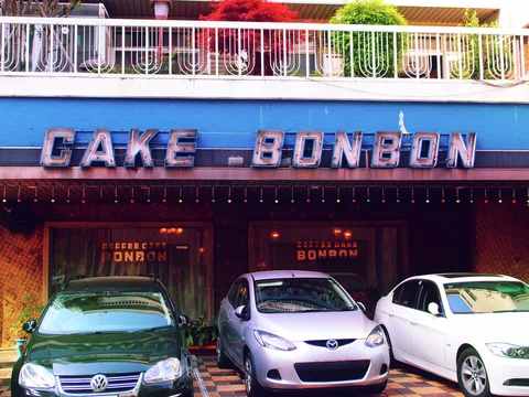 Three cars park outside a cake shop beneath large lettering and a balcony lined with greenery.