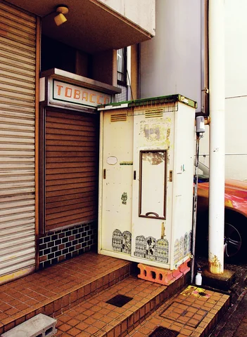 A weathered pale metal cabinet stands on bricks beside closed shop shutters and tiled steps.