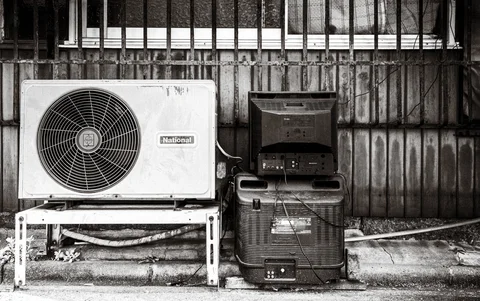 An air-conditioning unit and two stacked dark containers sit against a barred exterior wall.