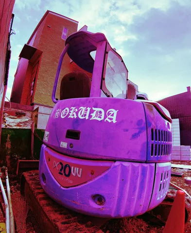 A compact purple-toned excavator sits beside a brick building and an orange traffic cone.