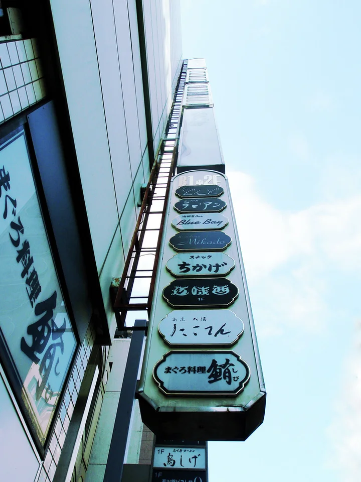 Stacked rounded business labels climb a narrow projecting directory against pale blue sky beside a building wall.