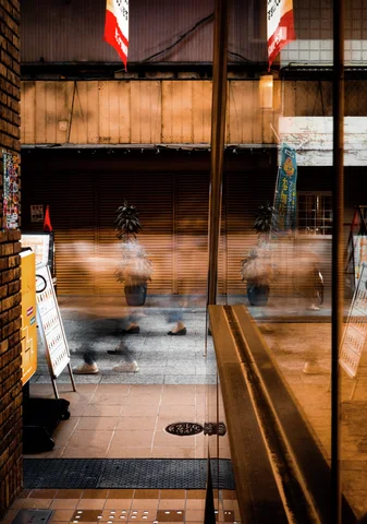 Blurred pedestrians pass beyond a glass entrance, reflected alongside warm lights and a wooden interior railing.