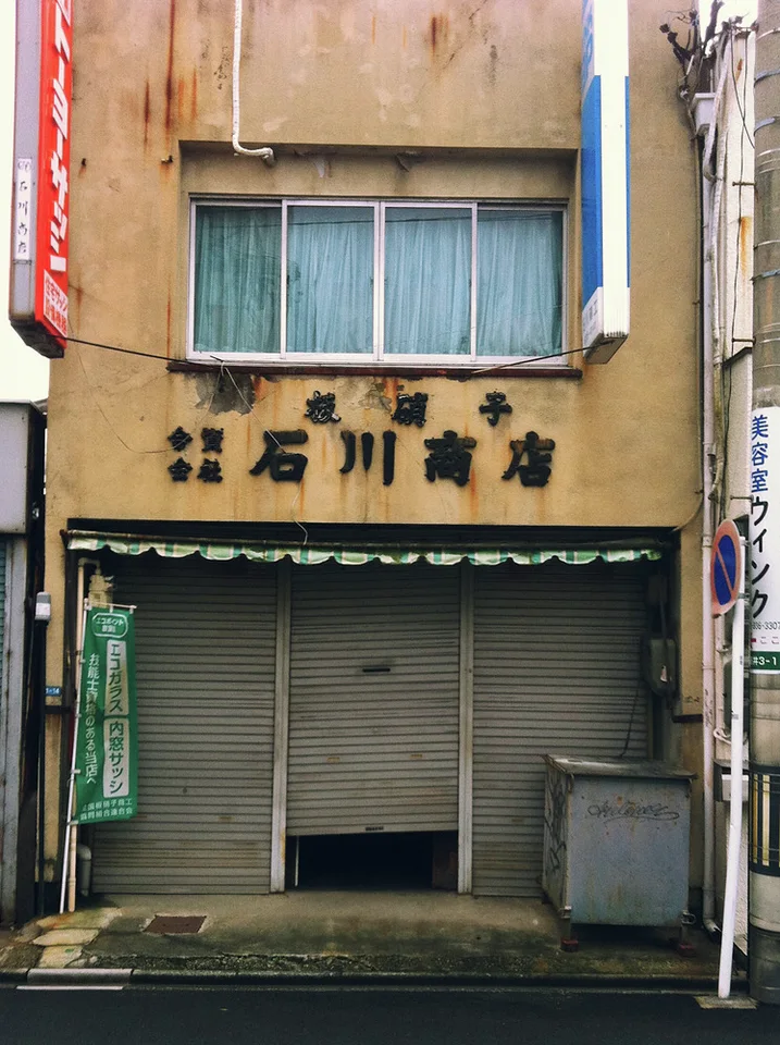Closed gray shutters sit beneath a faded tan facade with blue windows and dark lettering.