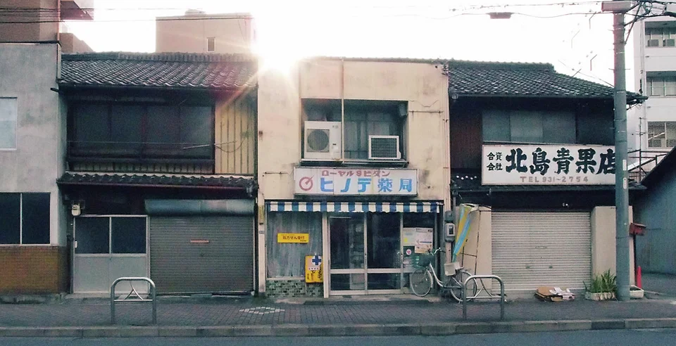 Sunlight flares above low tiled-roof shops, including a narrow pharmacy and closed bookshop.