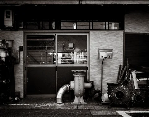 Large metal pipes and round industrial parts stand outside glass workshop doors in monochrome.