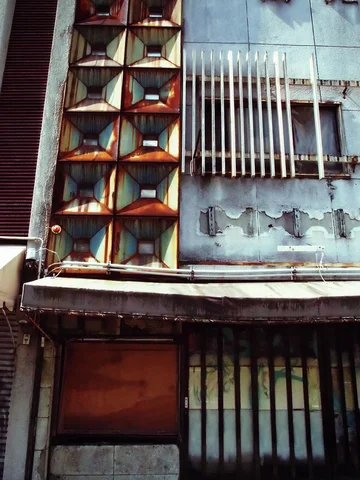 Geometric metal panels and a barred window sit above a weathered narrow storefront with peeling pale siding.