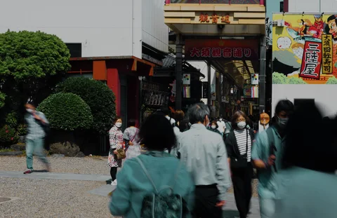 Pedestrians gather outside a covered shopping passage beside rounded shrubs and a colorful wall poster.