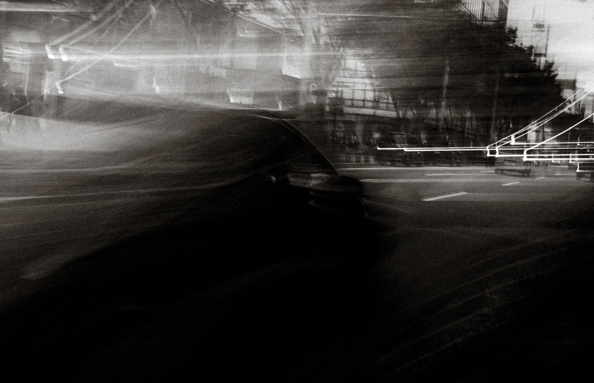 A blurred dark vehicle crosses a black and white street scene beneath streaked buildings and bright railings.