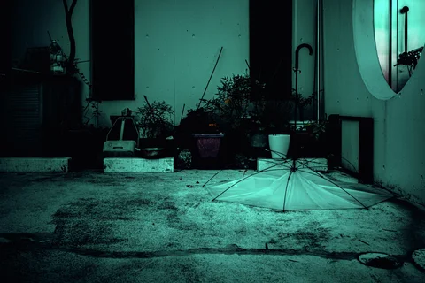 An open pale umbrella rests on turquoise-toned concrete beside potted plants and exterior pipes.