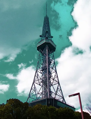 A steel lattice tower with a broad observation section rises above treetops beneath turquoise sky and white clouds.