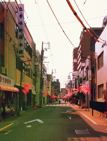 A narrow shopping street stretches between low storefronts beneath wires and hanging pink decorations.