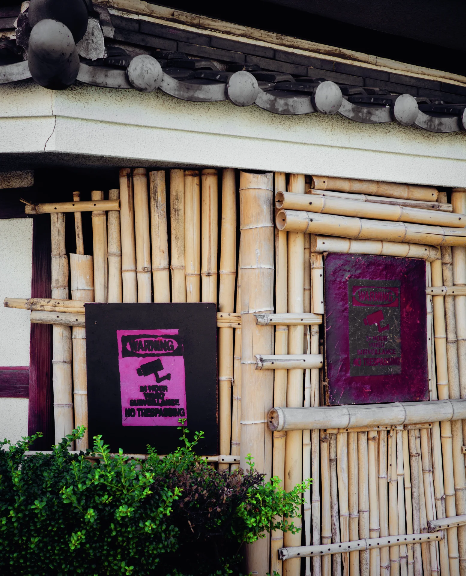 Vertical bamboo cladding surrounds dark framed notices beneath a gray tiled roof and leafy shrubs.