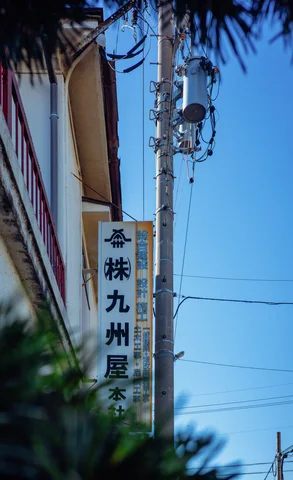 A vertical Japanese sign hangs beside a utility pole against blue sky framed by blurred foliage.