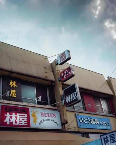 Multiple projecting shop signs angle outward from a pale building beneath a cloudy sky.