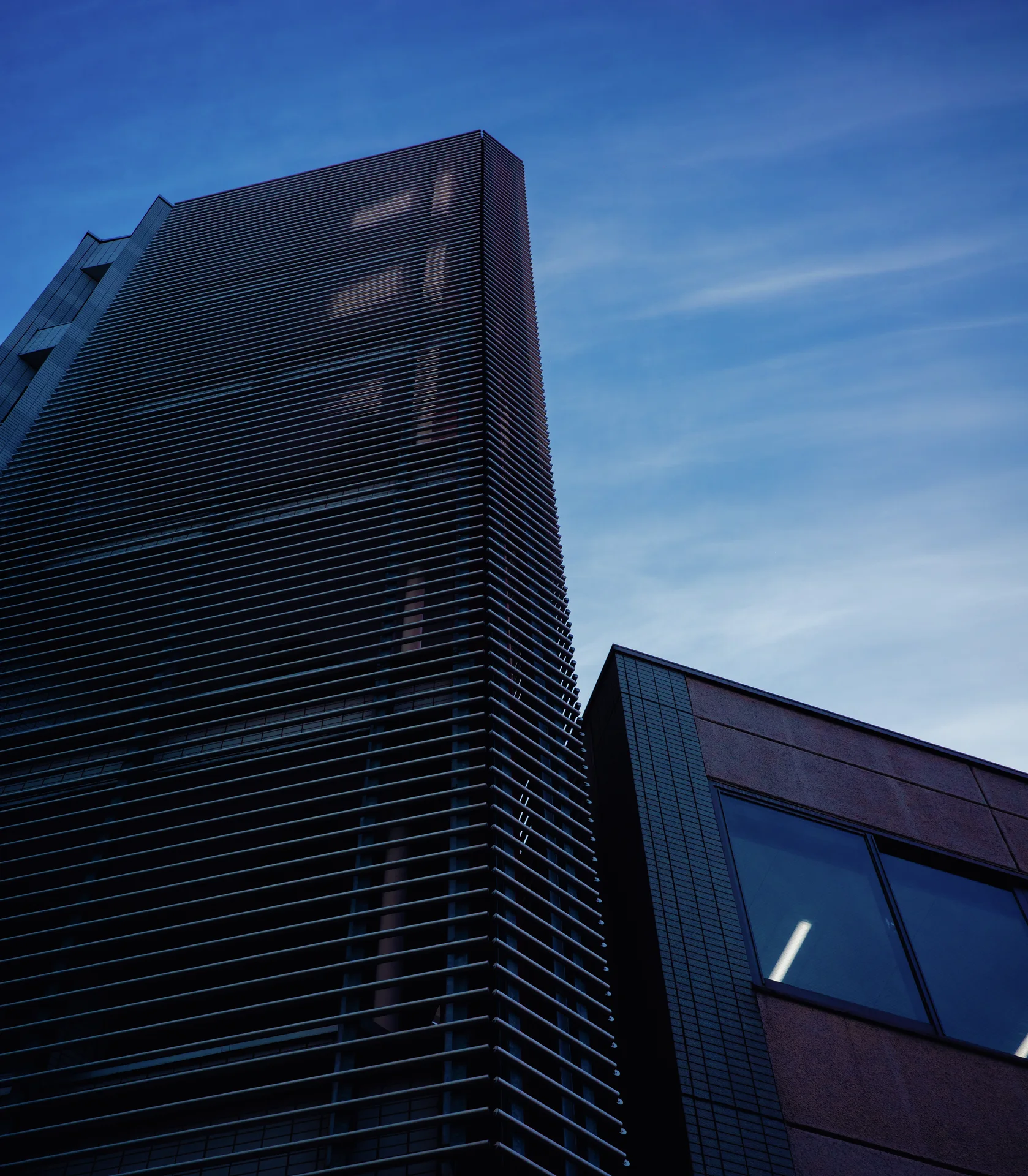 A tall dark building with dense horizontal louvers rises beside a lower structure against blue streaked sky.