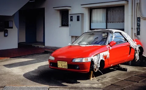 A red car rests on supports with missing wheels and torn covering across its roof.