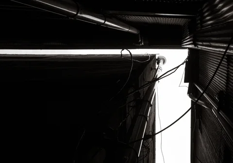 A narrow bright gap separates dark corrugated walls crossed by pipes and loose cables in black and white.
