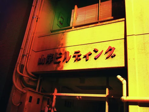 Raised red Japanese lettering crosses a yellow building facade crowded with exposed pipes and a small upstairs window.