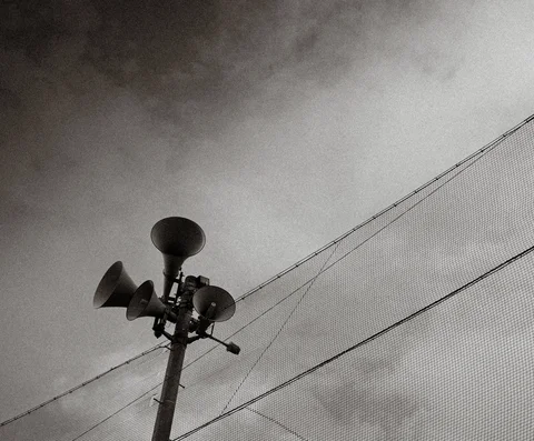 Horn loudspeakers rise beside overhead wires and tall netting against a monochrome sky.