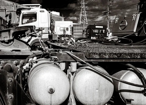 Round metal tanks and connecting hoses fill the foreground of a truck yard in black and white.