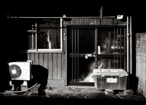 An air-conditioning unit and storage box flank a shadowed slatted house entrance in black and white.