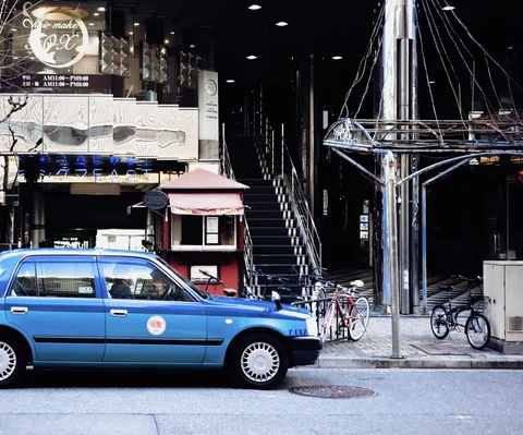 A blue taxi stands before a dark entrance with exterior stairs, parked bicycles, and a small red booth.