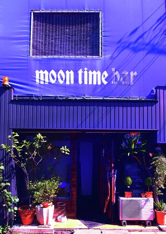 White Moon Time Bar lettering marks a deep blue facade above a dark entrance surrounded by potted plants.
