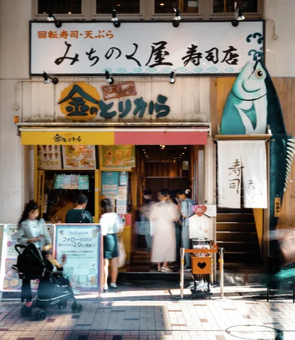 People gather around a small restaurant entrance beneath stacked signs and a tall blue fish graphic.