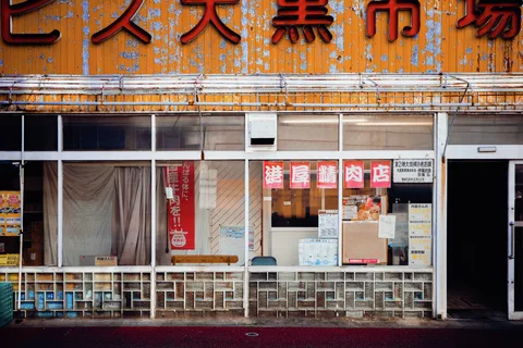 Large red Japanese characters crown a weathered orange storefront above broad glass windows and white railings.